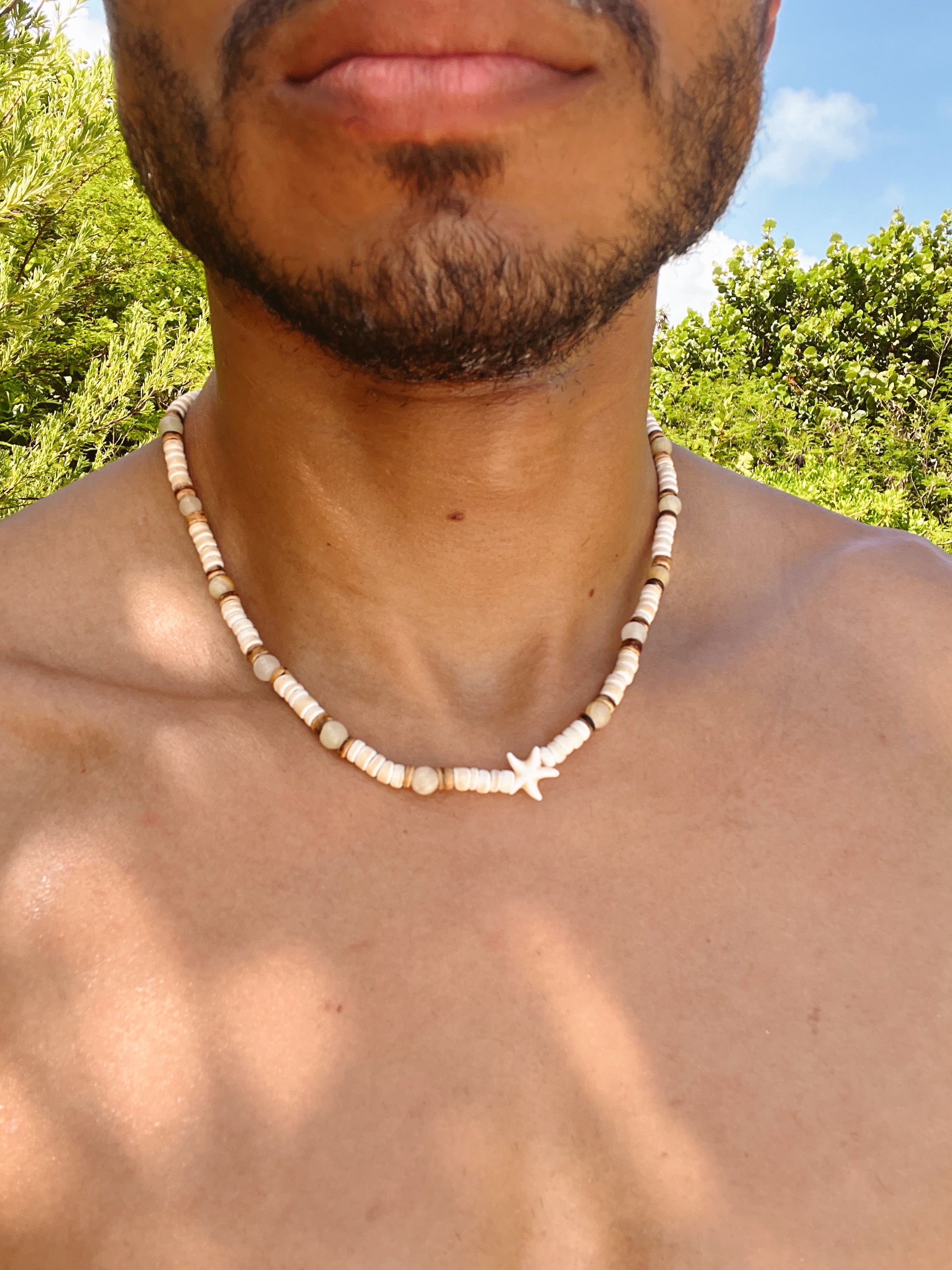 Man wearing a beaded necklace outdoors with greenery in the background