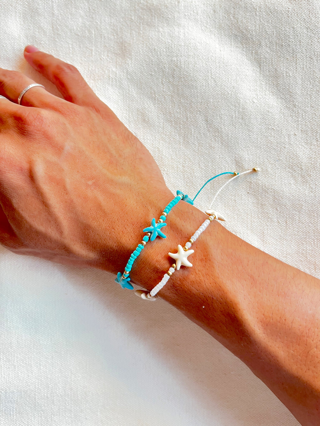 Hand wearing two bracelets, one turquoise with starfish charms and one white with starfish charms, on a white background.