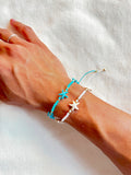 Hand wearing two bracelets, one turquoise with starfish charms and one white with starfish charms, on a white background.