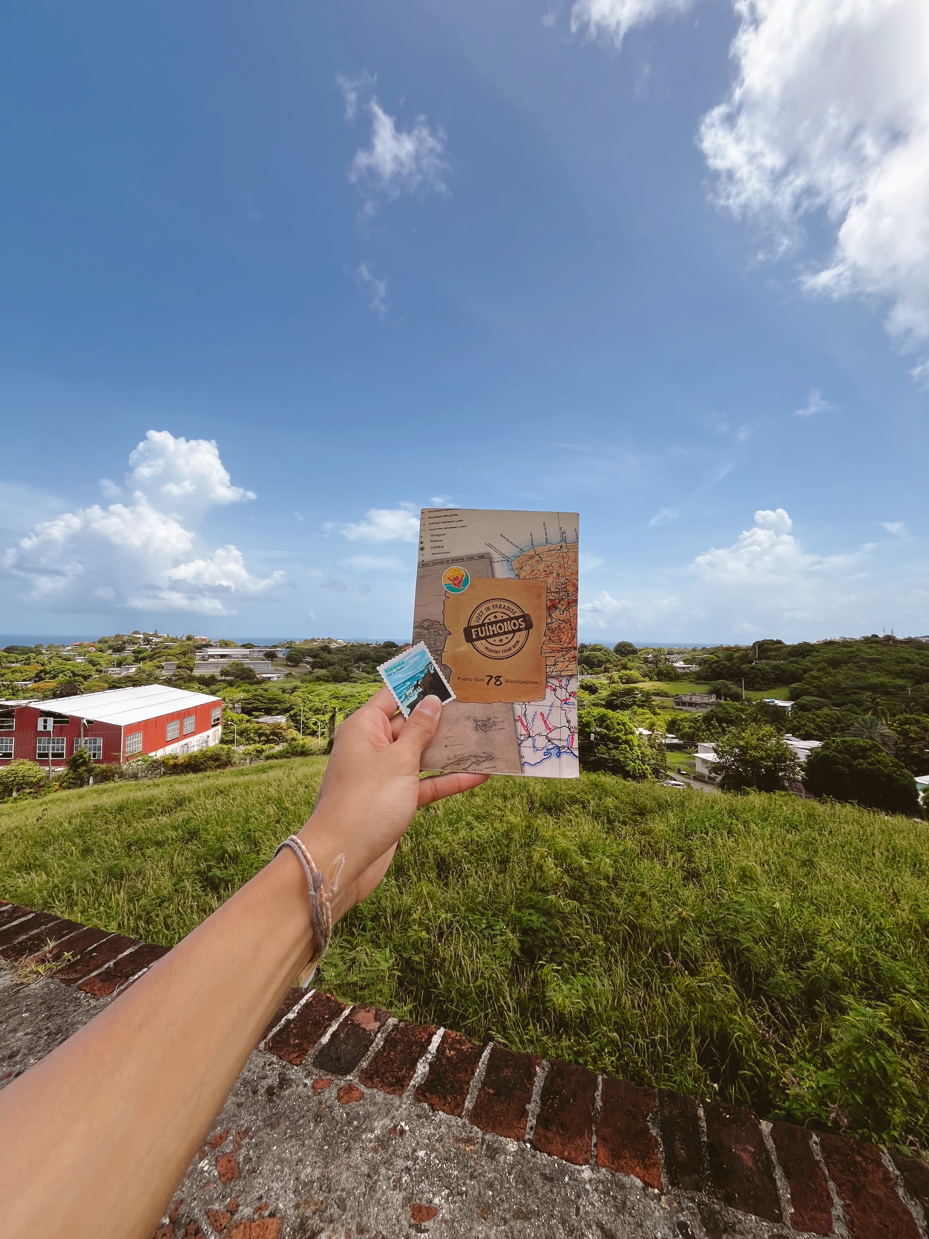 Hand holding a stamp book against a scenic backdrop with greenery and blue sky.